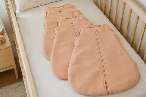 Trois gigoteuses bébé en coton abricot avec fermeture éclair à double sens, posées sur un lit clair.